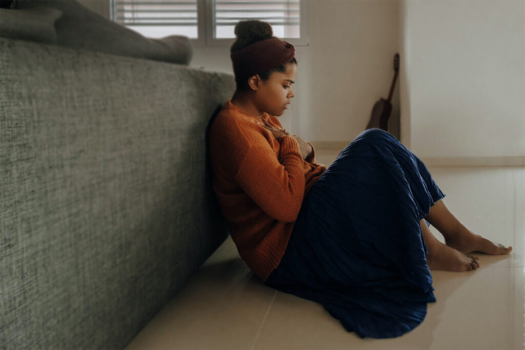 woman wearing orange longsleeves and a blue long skirt sitting on the floor beside the gray couch